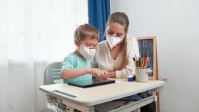 Mother with little son wearing protective medical respirator masks using tablet computer. Doing homework and studying at home during lockdown and self isolation. Remote school concept.