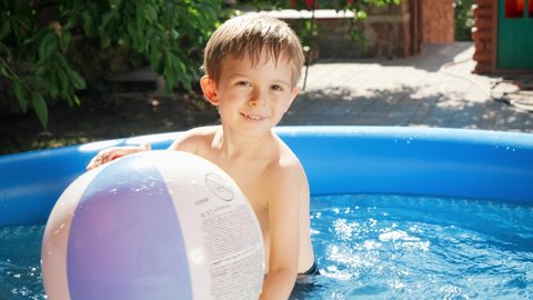 Happy smiling little boy playin with big colorful beach ball in inflatable swimming pool at house backyard garden. Concept of happy and cheerful summer holidays and vacation.