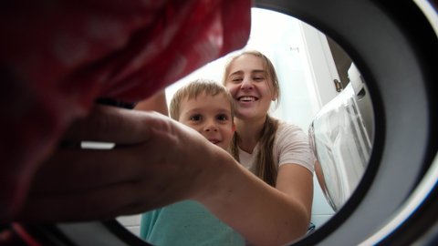 Smiling boy with young mother taking clean clothes out of washing machine in bathroom. Child helping parents washing clothes and doink chores.