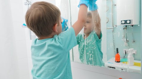 Little boy wearing rubber protective gloves while cleaning and washing bathroom mirrors with chemical detergent. Having fun while doing housework and home cleanup.