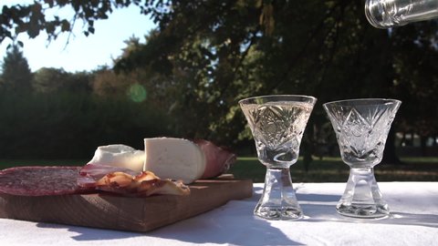 Pouring Rakija Brandy in Small Glasses on Picnic Table With Cheese, Sausage and Bacon. Traditional Refection in Balkans, Slow Motion