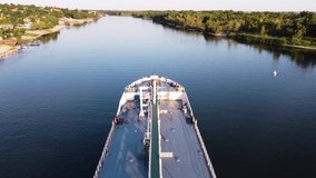 A large white cargo ship is anchored in the water area of the river. Footage. Aerial view of a ship in a river against the background of the green city on a summer sunny day.