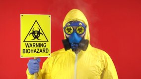 Show Signs of Warning Biohazard. A Man in a Protective Yellow suit and in a protective mask with a banner. Hazard Concept, Ecology Problem. Studio Footage, Red Background, 4K.
