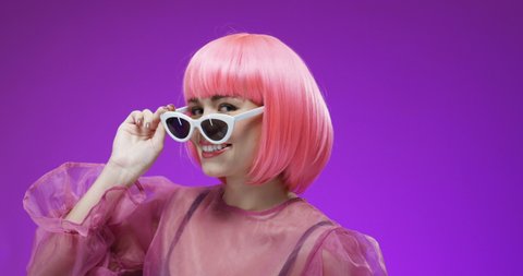 Portrait of pretty extraordinary young Caucasian woman in pink wig taking off fancy sunglasses and blinking to camera. Close up of happy cute female model smiling on bright background. Barbie concept.