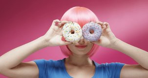 Close up of joyful pretty woman in pink wig smiling cheerfully and holding donuts in hands. Portrait of beautiful happy girl dancing with sweets on colorful background. Barbie doll look concept.