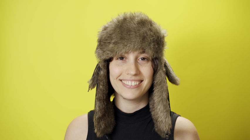 Happy young woman with fur trapper winter hat looks and smiles at camera. Cold weather conditions or fashion shoot. Beautiful and attractive female with fun and joyful look on yellow background