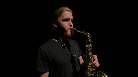Camera rotates in slow motion around young man with long hair playing the saxophone. Musician enjoys playing a wind instrument in the dark. Isolated on black background with studio light. Close up.