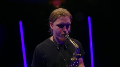 Camera rotates around a young man playing the saxophone. The musician performs in a dark studio amidst bright neon lights. Close up. Slow motion.