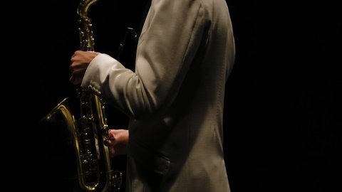 Saxophonist in a white suit performs in the dark. Jazzman with saxophone on black studio background with backlight. Torso and hands of a musician close up. Orbital shot. Slow motion.
