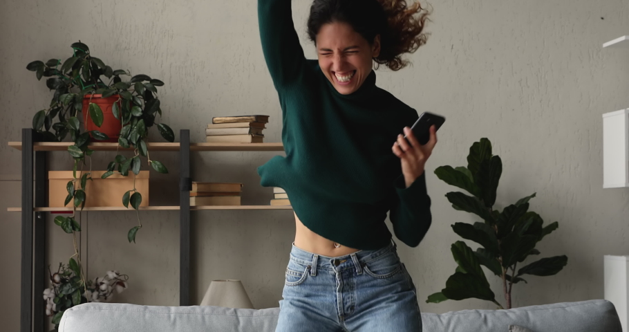 Woman sit on sofa check sms on smartphone read great news feels excited celebrate online lottery betting win scream with joy jumping look crazy, unbelievable happy. Achievement, moment of luck concept Royalty-Free Stock Footage #1065694543