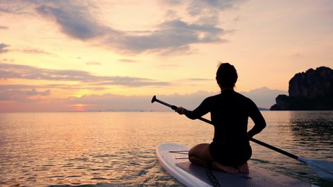Silhouette of Confidence Asian woman sit on sup board with paddleboarding passing beautiful sea with twilight sky at summer sunset. Female enjoy outdoor lifestyle and water sport on holiday vacation