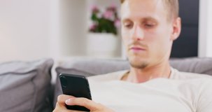 Happy Young Man Uses Smartphone while Sitting on a Sofa at Home. Man Browses Through Internet, Watches Videos and Uses Social Networks at Home.