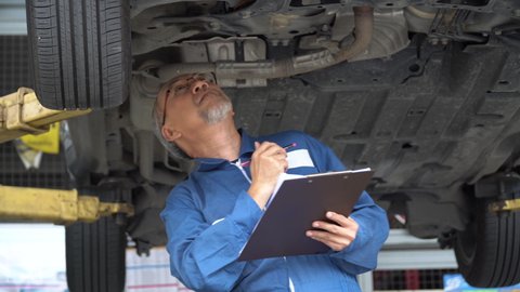 asian mechanic senior man holding clip board checking list to brake ,tyre, undercarriage of car in workshop at auto car repair service center with lift. car engineer old man inspection vehicle details