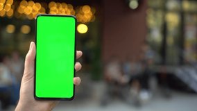 Close up hands woman holding phone with vertical green screen on busy street background pavement scrolling pages swiping surfing internet technology chroma key message