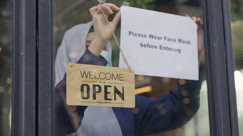 Young Asian Muslim woman in hijab wearing protective face mask, attach the sign on the shop front door to remind customer wear the face mask before entering for prevent the spread of coronavirus