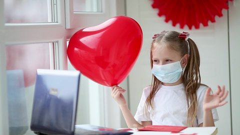 Happy little girl in a medical mask waves her hand, on the Internet celebrates Valentine's Day during quarantine, self-isolation. The child congratulates his friends via the Internet on February 14.