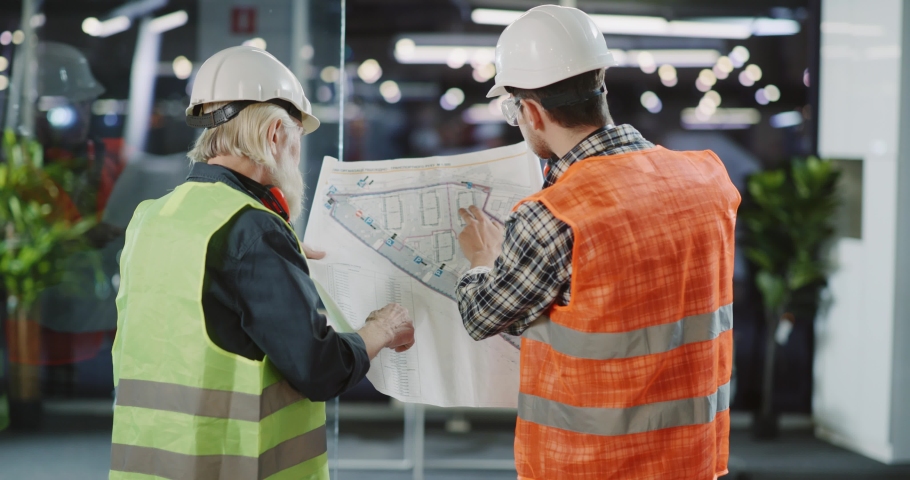Cooperative couple of multi-age workers wearing bodyvests and protective helmets inspecting construction plan of new enterprize company building. | Shutterstock HD Video #1065717943