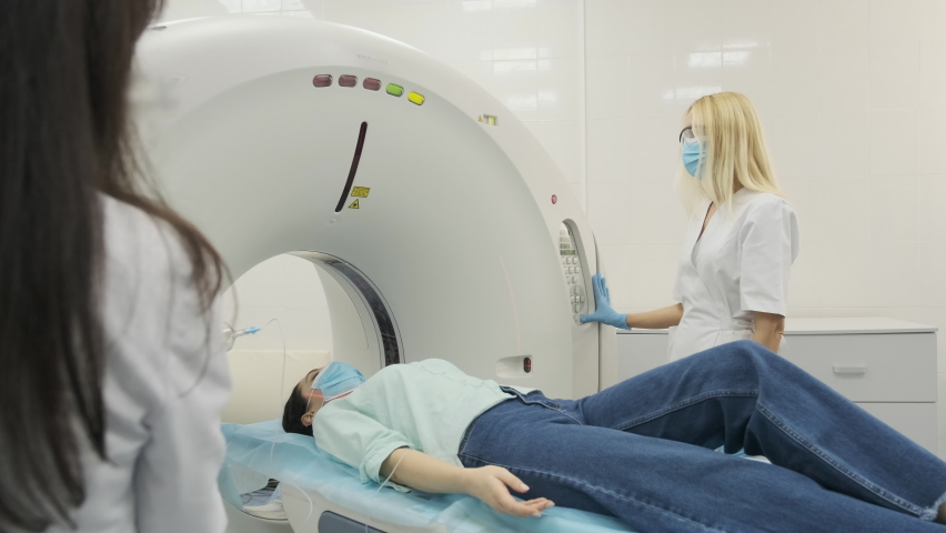 Female patient is undergoing CT or MRI scan under supervision of two qualified radiologists in modern medical clinic. Patient lying on a CT scan bed, in protective mask moving inside the machine. | Shutterstock HD Video #1065718225