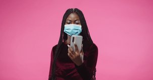 Portrait of joyful African American beautiful young woman in medical mask standing on rosy pink background wall and videochatting speaking on video call online on smartphone device