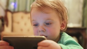A charming European baby with a new smartphone watches interesting videos or cartoons while lying on the floor. Close-up of a little boy with a phone resting at home. Selective focus
