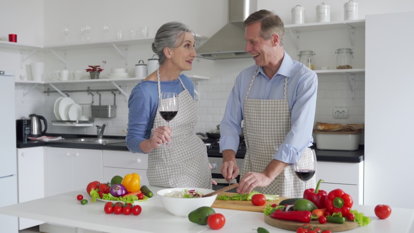 Happy old 50s couple wearing aprons having fun drinking wine talking, preparing meal in kitchen at home. Smiling senior mid aged husband and wife embracing, laughing, cooking, celebrating together. Royalty-Free Stock Footage #1065722650