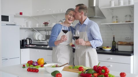 Happy old 50s couple wearing aprons having fun drinking wine talking, preparing meal in kitchen at home. Smiling senior mid aged husband and wife embracing, laughing, cooking, celebrating together.