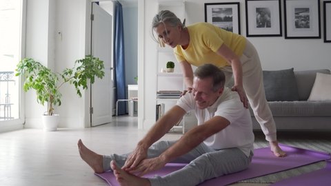 Fit active retired middle aged wife helping senior husband doing stretching exercise at home. Happy healthy older senior 60s couple enjoying fitness sport training workout together in apartment.