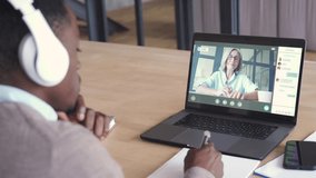 African business man or black male student wearing headphones conference video calling, watching webinar online, social distance learning or working using laptop at home office, over shoulder view.