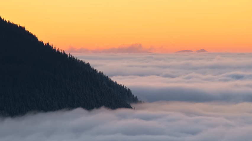 Time lapse of distant dark mountain hills surrounded with white fast moving foggy clouds at sunrise.