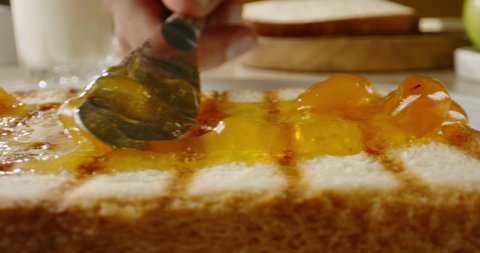 Preparing a sweet tasty sandwich for breakfast. Knife spreading apricot or peach jam on piece of fresh toast, close up 4k footage.