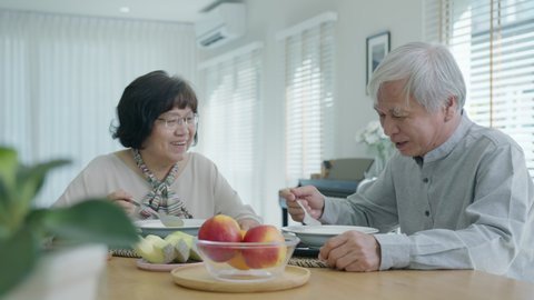 Attractive young senior asian citizen couple happy sit and talk, eat soup for healthy nutrition breakfast meal on dining table at home in morning in routine lifestyle in old asian people concept.