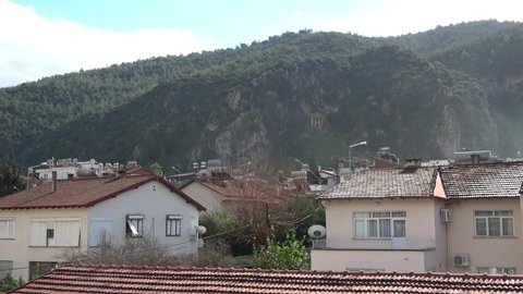Fethiye, Turkey - 13th of January 2021: 4K Lycian rock tomb viewed on hill behind red tiled roofs of old houses
