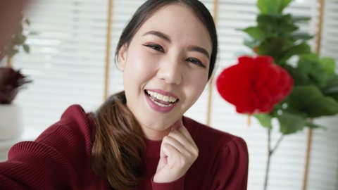 Point of view selfie videocall shot of attractive beautiful young asian lady in red chinese new year celebration outfit happy smile talk and looking to camera mobile phone wishing for lunar festival.