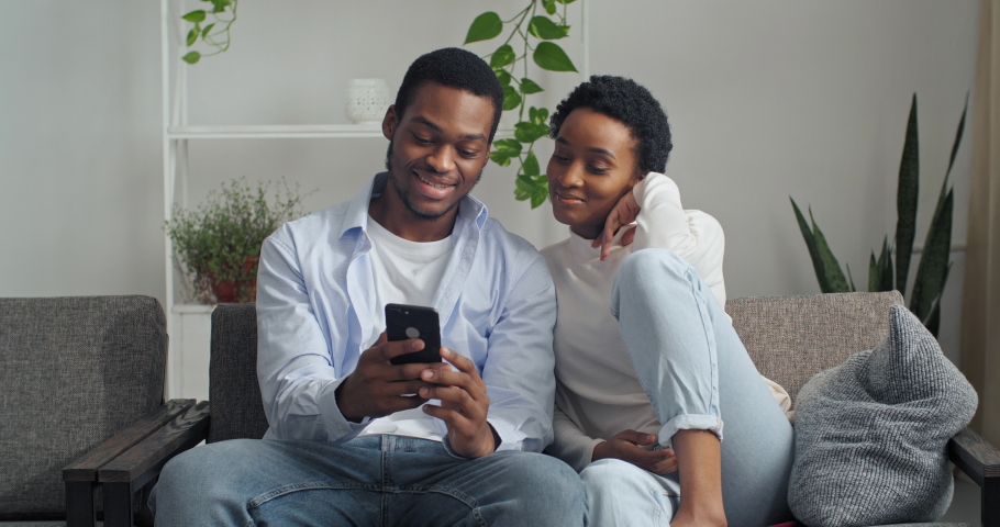 Afro american couple man and woman sitting together at home looking at phone screen using mobile device gadget to search for booking tickets for vacations choosing hotel on internet discussing talking Royalty-Free Stock Footage #1065737707