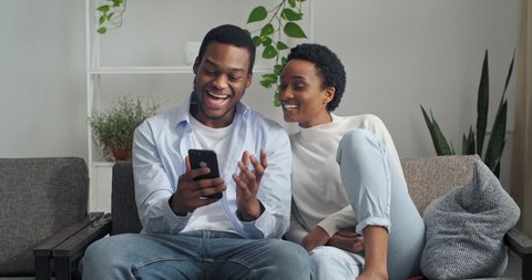 Afro american couple man and woman sitting together at home looking at phone screen using mobile device gadget to search for booking tickets for vacations choosing hotel on internet discussing talking