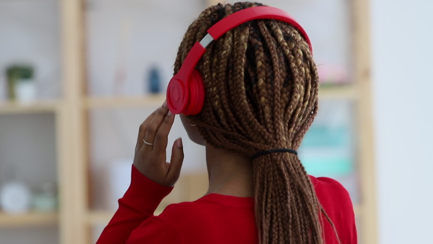 Beautiful young woman is listening to music and dancing in apartment room. Front view of African American female enjoying sound and moving body in light interior. Pretty millennial lady using