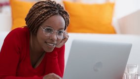 Young woman lying on sofa, looking at laptop, typing and browsing internet. Relaxing on bed, watching searching internet movie. happy african american young woman in glasses