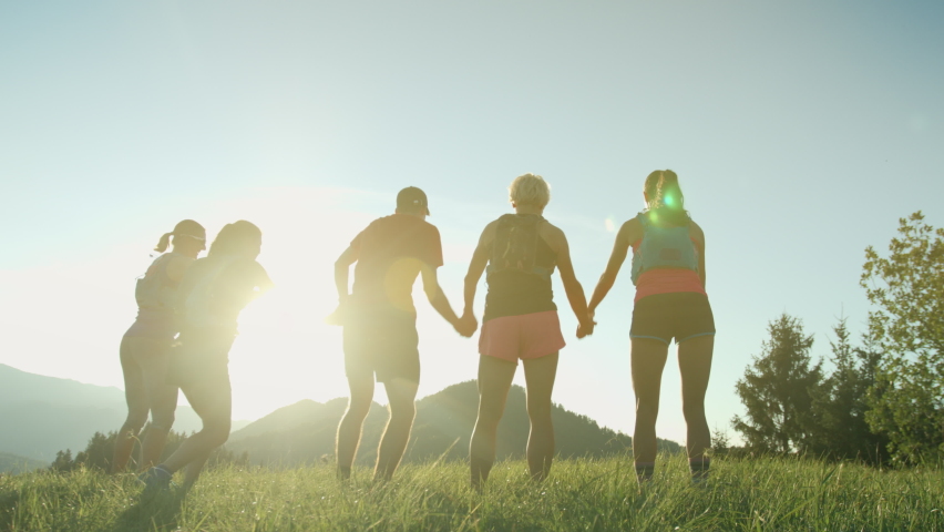 A team of runners jumps in the air together on the top of the hill with mountain view in a beautiful golden sunset light. Runners holding their hands and together reach the goal. Royalty-Free Stock Footage #1065743107