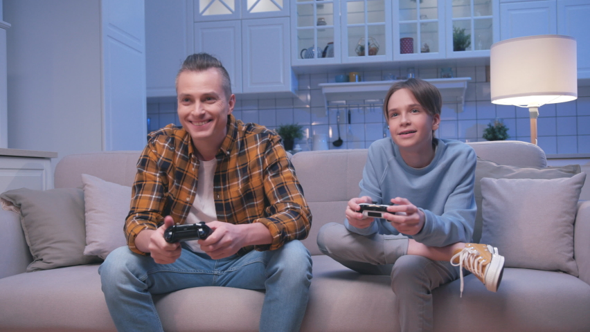 Action Gamers Adult Man and Guy Sitting on Sofa and Playing Video Games on Digital Console. Two Male Players Gaming in Controlling Joysticks for Videogame. Having Fun Focused on Gaming in Living Room | Shutterstock HD Video #1065744397