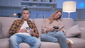 Action Gamers Young Man and Woman Sitting on Sofa and Playing Video Games on Digital Console. Two Players Gaming in Controlling Joysticks for Videogame. Having Fun Focused on Gaming in Living Room 4k