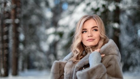 Portrait of fashionable feminine posing in gray hood fur coat smiling outdoor at winter nature park. Adorable blonde lady playing warm luxury clothes in forest. Shot on RED Raven 4k Cinema Camera