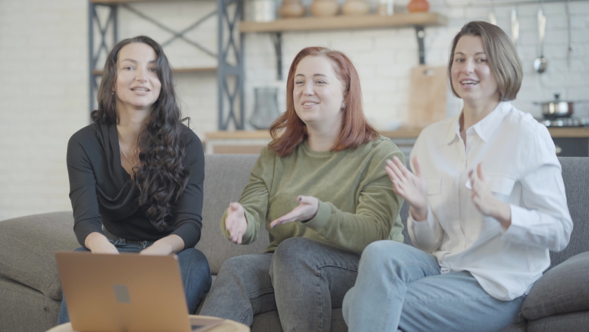 Three joyful Caucasian women watching comedy show on laptop laughing and clapping. Positive carefree friends having fun enjoying leisure indoors. Resting and relaxation concept.