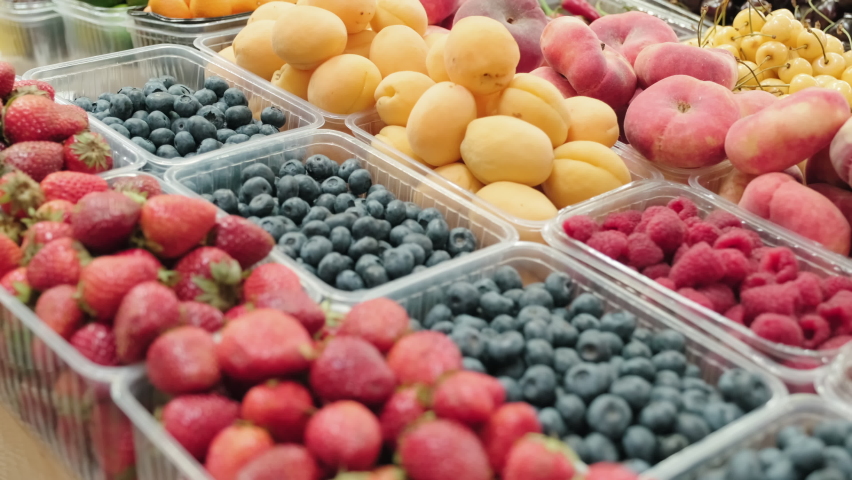 Handheld tracking close up of tasty strawberries, blueberries, raspberries, peaches and apricots in disposable plastic food containers for sale at market