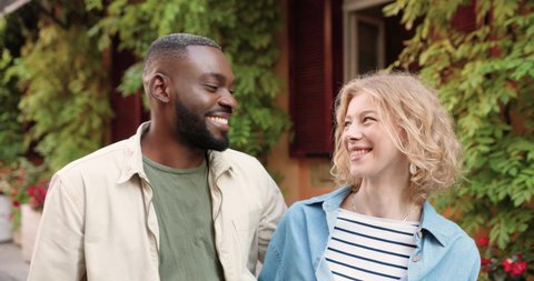 Portrait of young happy mixed-races couple standing outdoors, hugging and looking at camera. Stylish multi ethnic male and female smiling, embracing and posing in town. Close up.