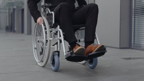 Crop view of disabled male person in stylish shoes and suit going on wheel chair at street. Successful male person riding to work near office building. Concept of motivation, grid
