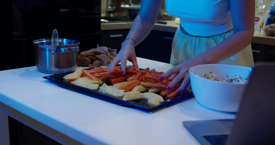 A modern, stylish and luxurious kitchen with light blue ambiance. A baking tray with fresh vegetables. A woman prepares healthy food in the kitchen. A baking tray with potatoes and carrots.
