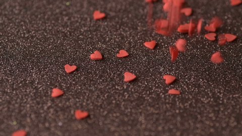 Red scarlet heart shaped sugar confetti piece lies, more fall down cover scatter light brown shiny surface slow motion. Romantic love, affection or Saint Valentine's Day preparation design concept