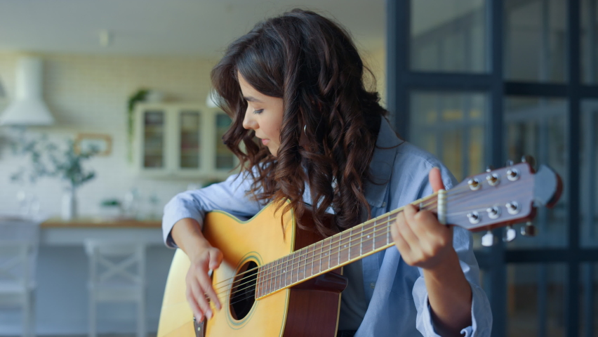 Excited girl playing acoustic guitar in living room. Happy musician playing chords on string instrument. Positive guitarist moving head during song. Musician performing musical composition on guitar | Shutterstock HD Video #1065765808