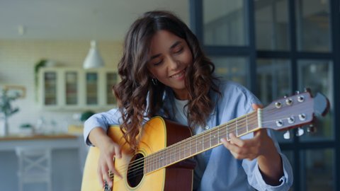 Excited girl playing acoustic guitar in living room. Happy musician playing chords on string instrument. Positive guitarist moving head during song. Musician performing musical composition on guitar
