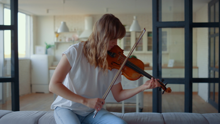 Teenage girl playing violin in living room. Female violinist playing chords on musical instrument at home. Inspired musician pressing strings of violin. Young woman sitting on sofa Royalty-Free Stock Footage #1065765949